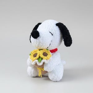 Può includere: Un cane di peluche bianco fatto a mano con orecchie nere e naso nero, con un collare rosso. Il cane tiene in mano un cestino di girasoli gialli con foglie verdi. Il peluche è seduto su una superficie bianca.