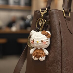 May include: A handmade crochet Hello Kitty keychain in white and brown, hanging from a brown leather handbag. The keychain features a white head with a brown bow, brown overalls, and a yellow nose. The bag has a gold-colored clasp.