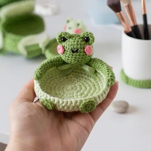 Op de afbeelding: Een handgemaakte gehaakte kikker-vormige sieradenschaal in groentinten, met roze wangen en een klein groen strikje. De kikker heeft een rond lichaam en wordt in de hand van een persoon gehouden. Andere gehaakte kikker-items staan op de achtergrond.