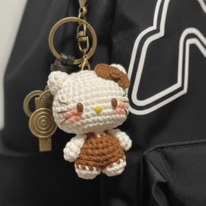 May include: A handmade crochet keychain featuring a white and brown Hello Kitty character. The keychain has a gold-colored clasp and is attached to a black backpack. The character has a brown bow, pink cheeks, and a yellow nose.