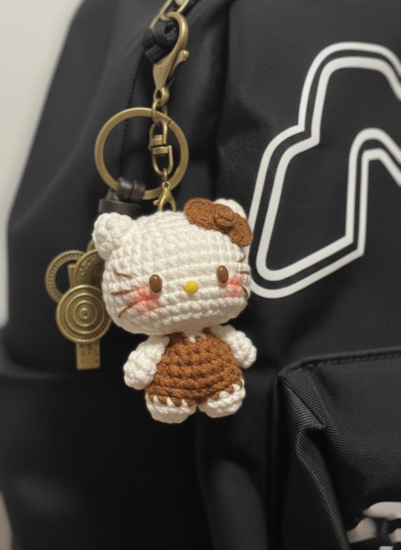 May include: A handmade crochet keychain featuring a white and brown Hello Kitty character. The keychain has a gold-colored clasp and is attached to a black backpack. The character has a brown bow, pink cheeks, and a yellow nose.