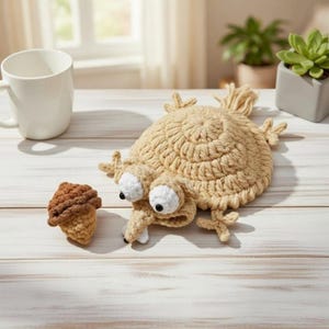 May include: A whimsical, handmade crochet creature with large, white-rimmed eyes and a long snout. The tan-colored body is round with small appendages. Beside it is a small, brown and tan crochet acorn. The items are on a light wood surface.