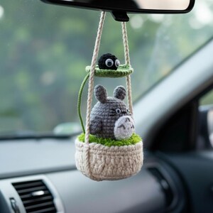 Może przedstawiać: Ręcznie robiony szydełkowy urok do samochodu z szarą postacią podobną do Totoro w beżowym koszu z zielonymi akcentami, zawieszony na lusterku wstecznym. Mniejsza czarna postać siedzi powyżej na zielonej platformie.