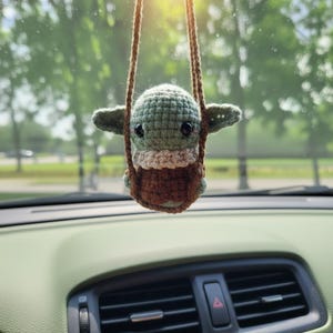 May include: A crocheted Baby Yoda car charm, suspended from a brown string. The charm has a green head, tan neck, and brown body, sitting in a brown crocheted swing. The background shows a car dashboard and a blurred outdoor scene.