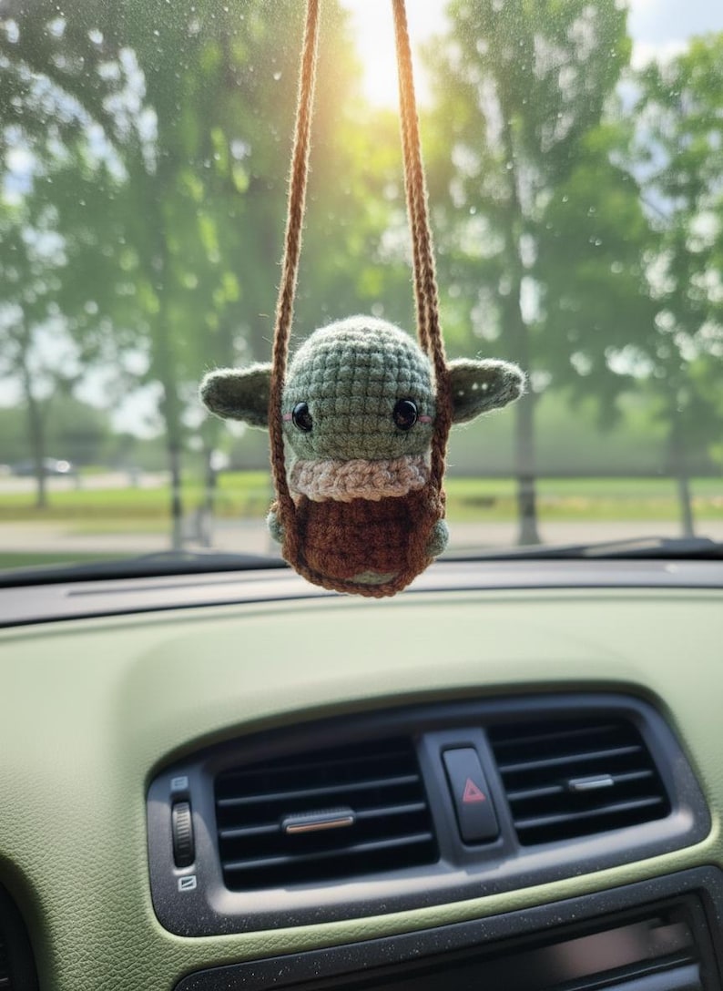 May include: A crocheted Baby Yoda car charm, suspended from a brown string. The charm has a green head, tan neck, and brown body, sitting in a brown crocheted swing. The background shows a car dashboard and a blurred outdoor scene.