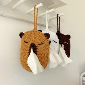 May include: Three crocheted tissue box covers shaped like bears, in brown, white, and dark brown. Each bear has closed eyes and a tissue emerging from its nose. The covers hang from a white rack.