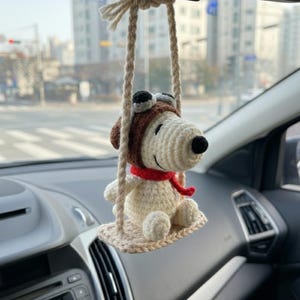 Può includere: Un ciondolo per auto di Snoopy all'uncinetto appeso allo specchietto retrovisore di un'auto. Il ciondolo raffigura Snoopy con un cappello da aviatore marrone, occhiali neri e una sciarpa rossa, seduto su un'altalena beige.