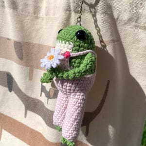 Könnte beinhalten: Ein handgefertigter Frosch-Schlüsselanhänger aus Häkelarbeit. Der grüne Frosch hat schwarze Augen, trägt eine rosa Latzhose und hält eine weiße Gänseblümchen. Der Schlüsselanhänger ist an einem silbernen Ring befestigt.