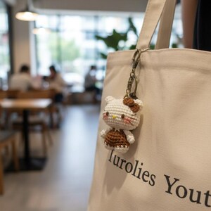 May include: A crocheted Hello Kitty keychain with a brown bow and dress, attached to a beige tote bag. The bag has the text "Turolies Youth" printed on it. The background shows a blurred cafe setting.