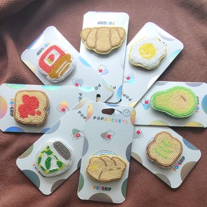 May include: A set of 10 colorful beaded phone grips with various designs, including avocado, egg, toast, and a variety of other food items. The grips are all labeled with the brand name "PopGrip" and "PopSockets".