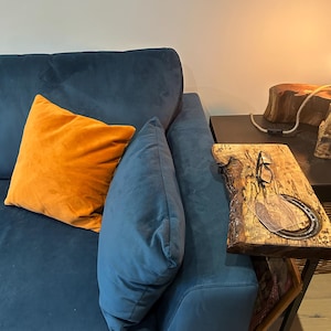 Puede incluir: Un sofá de terciopelo azul marino con un cojín naranja y otro azul. Una mesa auxiliar de madera con una herradura y gafas está al lado del sofá. La habitación está iluminada por una luz cálida.