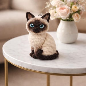 May include: A crocheted Siamese cat ornament with blue eyes, brown markings, and a cream body. The cat sits on a marble-topped table with gold legs. A vase of flowers is in the background.