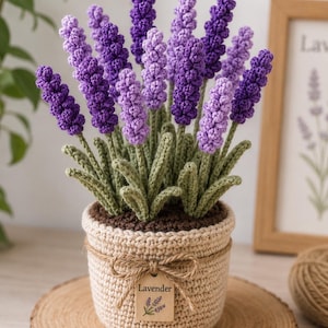 May include: A crocheted lavender plant in a beige pot. The lavender flowers are various shades of purple, with green stems and leaves. The pot is tied with twine and has a small tag that says "Lavender". The plant sits on a wooden slice.