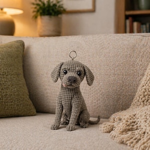 May include: A crocheted dog keychain in gray sits on a beige couch. The dog has floppy ears, a small tail, and a metal ring for attaching to keys. A green pillow and a beige throw blanket are also visible.