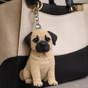 Könnte beinhalten: Ein gehäkelter Mops-Schlüsselanhänger in Beige und Schwarz, der an einer schwarzen und cremefarbenen Handtasche hängt. Der Mops hat dunkle Augen, eine schwarze Nase und Ohren. Die Handtasche hat schwarze Griffe und goldfarbene Beschläge.