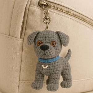 May include: A grey, crocheted dog keyring with a blue collar and a white chevron detail. The dog has brown eyes and is attached to a metal clasp. The keyring is hanging from a beige backpack.