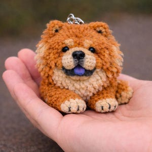 Könnte beinhalten: Ein handgefertigter Chow Chow-Hunde-Schlüsselanhänger. Das Plüschtier ist überwiegend orange mit helleren beigen Akzenten auf Gesicht und Pfoten. Es hat blaue Augen und eine lila Zunge. Der Schlüsselanhänger besteht aus einem weichen, strukturierten Material.