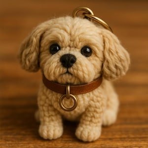 Puede incluir: Un llavero de cachorro beige hecho a mano, con un collar de cuero marrón y herrajes dorados. El peluche tiene ojos grandes y oscuros y una nariz negra. El llavero está diseñado para parecerse a un perro pequeño.
