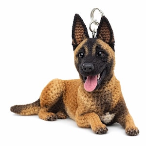May include: A crocheted dog keychain in a lying down position. The dog has brown and black fur, with a pink tongue sticking out. The keychain has a silver ring attached to the top.