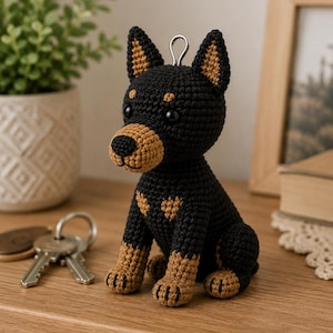 May include: A small, handcrafted crochet dog figurine featuring black yarn for the main body and tan accents on the muzzle, chest, ears, and paws. The figure has black plastic eyes, a stitched nose, and a silver metal keyring loop on top.