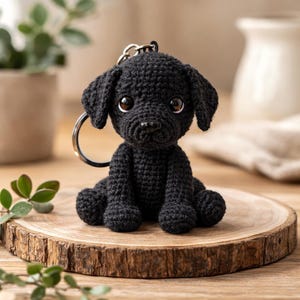 Könnte beinhalten: Ein schwarzer gehäkelter Welpen-Schlüsselanhänger mit großen, ausdrucksstarken Augen und einem silberfarbenen Schlüsselring. Der handgefertigte Hund sitzt auf einer Holzscheibe und zeigt seine detaillierte Handwerkskunst. Die Ohren des Welpen sind schlaff.