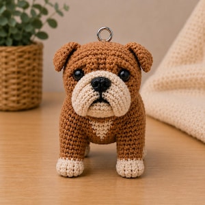Könnte beinhalten: Eine braun-cremefarbene Häkelfigur eines Bulldogs mit schwarzen Kunststoffaugen und einer schwarzen Nase. Der Hund hat einen kleinen Metallring am Kopf, der als Schlüsselanhänger oder Aufhänger dient. Die Häkelstruktur ist deutlich erkennbar.