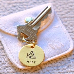 Peut inclure: Une clé dorée et un porte-clés rond avec un motif de maison et d'arbre. Le porte-clés porte les initiales "M & T" gravées sous la maison. La clé et le porte-clés sont sur une pochette blanche.