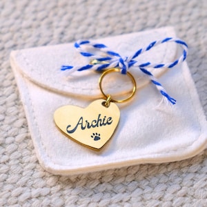 Peut inclure: Une étiquette dorée en forme de cœur pour animaux de compagnie avec le nom "Archie" et une empreinte de patte. L'étiquette est suspendue à un anneau doré et attachée à une pochette blanche nouée avec de la ficelle bleue et blanche. Accessoire pour animaux.