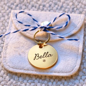 May include: A gold-coloured, round pet tag engraved with the name "Bella" and a small flower design. The tag hangs from a gold ring and is attached to a small, white pouch tied with blue and white twine.