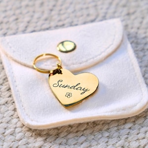 May include: A gold-toned heart-shaped metal tag with a circular ring attachment. The tag features the word Sunday in a cursive script font and a small flower icon engraved on the surface. It is placed on a small white fabric pouch with a gold snap.