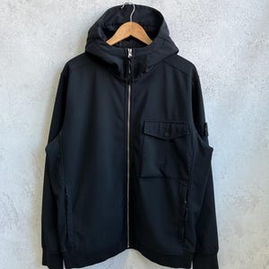 May include: A black hooded jacket with a full-length zipper and two front pockets. The jacket is hanging on a wooden hanger against a gray background. The jacket has a small logo on the left sleeve.