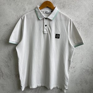 Może przedstawiać: Biała koszulka polo z jasnozielonym wykończeniem na kołnierzu i rękawach. Koszulka ma zapięcie na trzy guziki i małą czarno-zieloną naszywkę z logo na piersi. Koszulka wisi na drewnianym wieszaku.