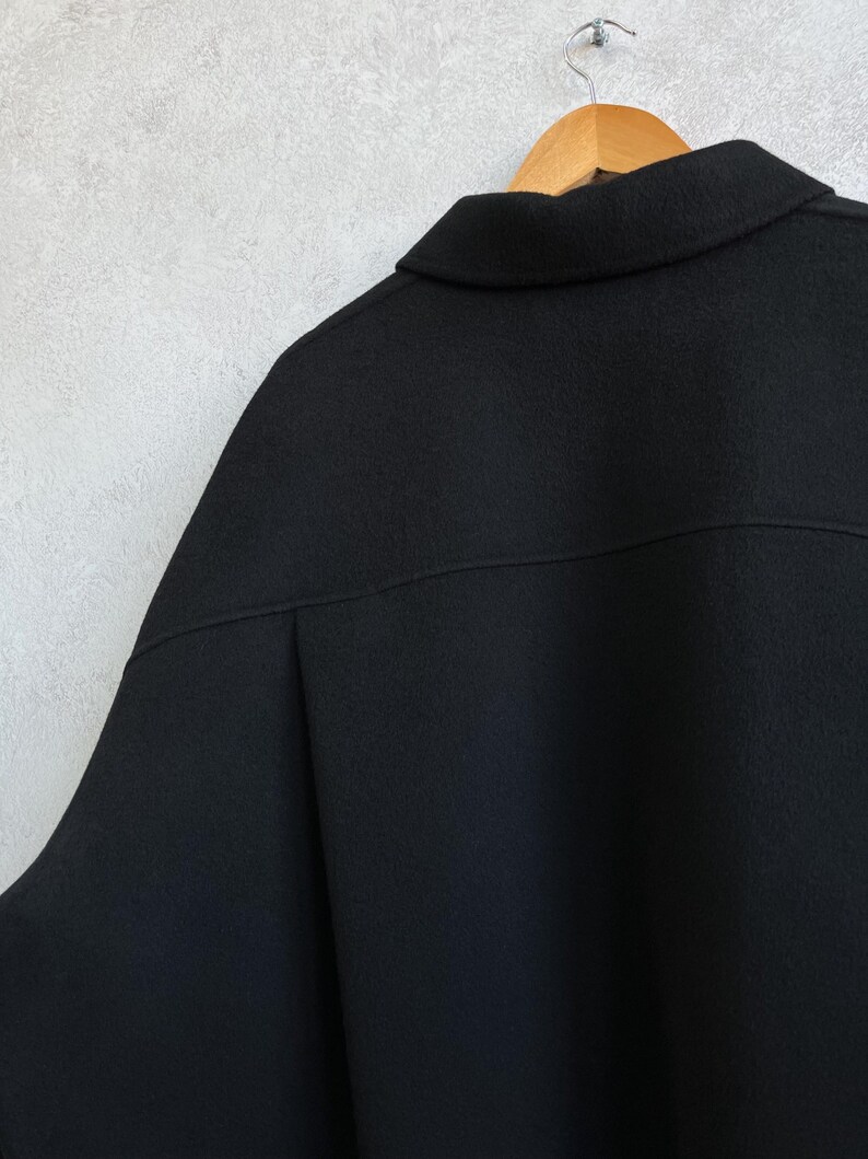 Pode incluir: Um casaco preto com gola, pendurado num cabide de madeira. O casaco &eacute; feito de um tecido escuro e liso. A gola est&aacute; dobrada e o casaco est&aacute; contra uma parede cinza claro.