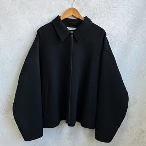 Pode incluir: Casaco preto com gola e fecho de correr. O casaco tem dois bolsos e a marca "Acne Studios" &eacute; vis&iacute;vel na etiqueta. O casaco est&aacute; pendurado num cabide de madeira.