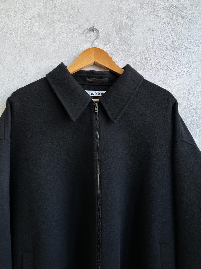 Pode incluir: Casaco preto com gola e fecho de correr. O casaco est&aacute; pendurado num cabide de madeira. O nome da marca "Acne Studios" &eacute; vis&iacute;vel na gola. O casaco &eacute; feito de um material espesso.