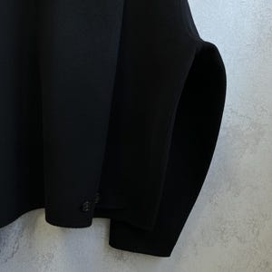 Pode incluir: Um close-up de um casaco preto. O casaco tem um design simples com gola redonda e dois bot&otilde;es na manga. O tecido parece ser um material liso e fosco. A silhueta do casaco &eacute; limpa e moderna.