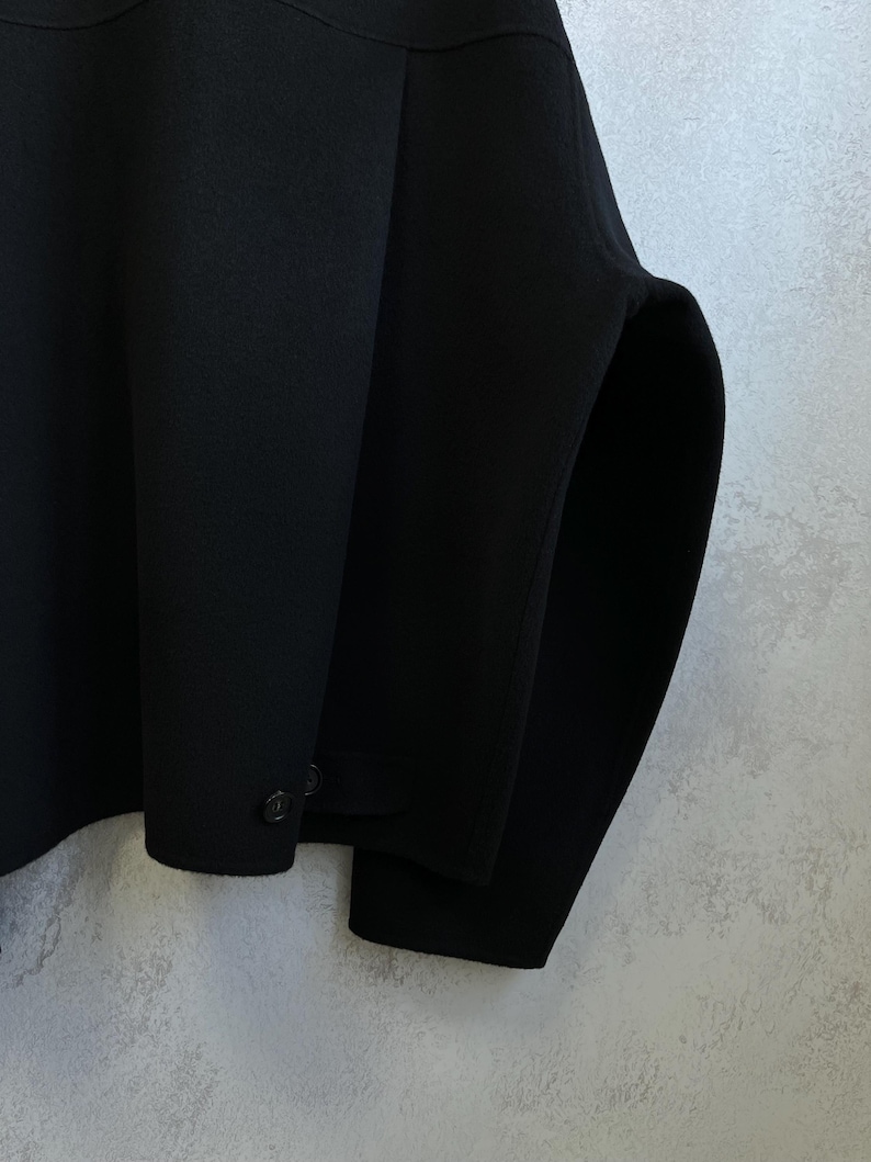 Pode incluir: Um close-up de um casaco preto. O casaco tem um design simples com gola redonda e dois bot&otilde;es na manga. O tecido parece ser um material liso e fosco. A silhueta do casaco &eacute; limpa e moderna.