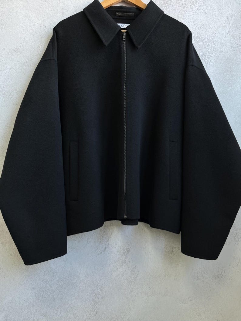 Pode incluir: Casaco preto com gola e z&iacute;per de comprimento total. O casaco tem dois bolsos frontais e mangas compridas. Est&aacute; pendurado num cabide de madeira.