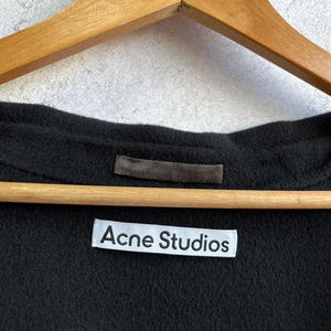 Pode incluir: Close-up de uma pe&ccedil;a de roupa preta com uma etiqueta branca que diz "Acne Studios". A pe&ccedil;a de roupa est&aacute; pendurada num cabide de madeira. O tecido parece ser um material macio e escuro.