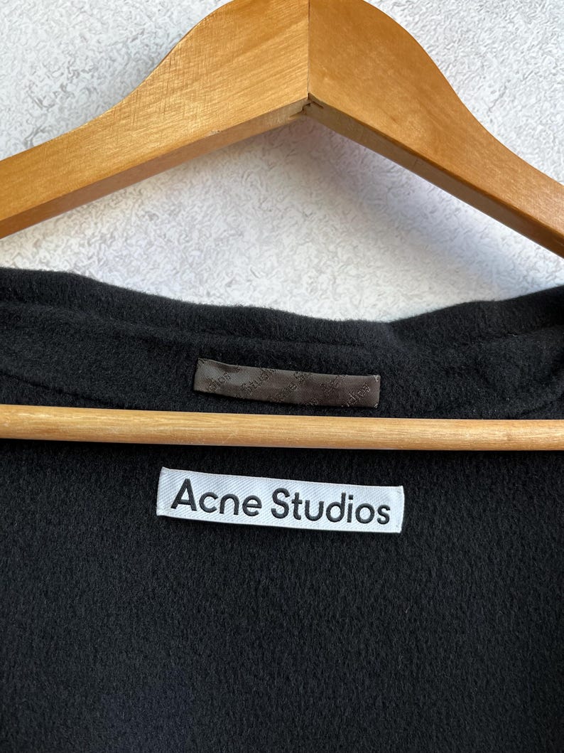 Pode incluir: Close-up de uma pe&ccedil;a de roupa preta com uma etiqueta branca que diz "Acne Studios". A pe&ccedil;a de roupa est&aacute; pendurada num cabide de madeira. O tecido parece ser um material macio e escuro.