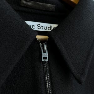 Pode incluir: Close-up de um casaco preto com gola e z&iacute;per prateado. A etiqueta diz "Acne Studios" em uma etiqueta branca. O tecido parece ser um material macio e tecido.