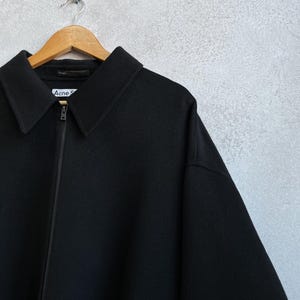 Pode incluir: Casaco preto com gola e fecho de correr, pendurado num cabide de madeira. O nome da marca "Acne S" &eacute; vis&iacute;vel na etiqueta. O casaco &eacute; feito de um material espesso.