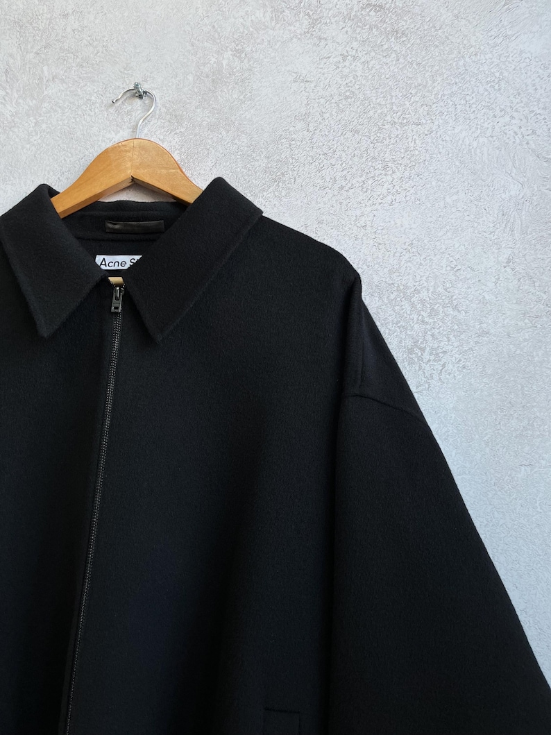 Pode incluir: Casaco preto com gola e fecho de correr, pendurado num cabide de madeira. O nome da marca "Acne S" &eacute; vis&iacute;vel na etiqueta. O casaco &eacute; feito de um material espesso.