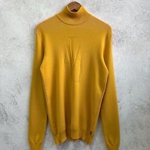 May include: Mustard yellow turtleneck jumper with long sleeves. The jumper has a raised design on the front. The garment is hanging on a wooden hanger against a textured, light grey background.