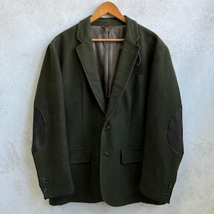 May include: Dark green blazer with brown elbow patches, two front pockets, and a notched lapel. The jacket is hanging on a wooden hanger against a light gray textured wall. The blazer has three buttons on each cuff.