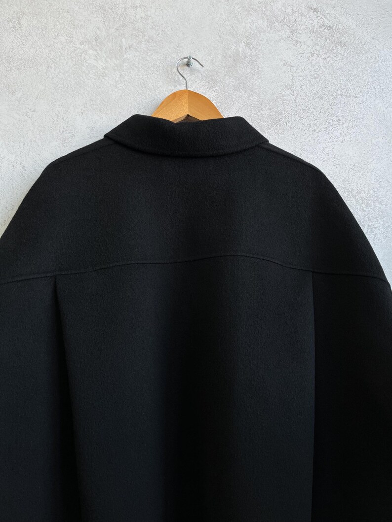 Pode incluir: Um casaco preto pendurado em um cabide de madeira contra uma parede cinza claro. O casaco tem uma gola larga e um design simples e cl&aacute;ssico. O tecido parece ser um material macio e tecido. A parte de tr&aacute;s do casaco &eacute; vis&iacute;vel, mostrando a constru&ccedil;&atilde;o sob medida.