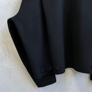 Pode incluir: Uma caneca de cer&acirc;mica branca com uma al&ccedil;a preta e o texto "But First, Coffee" em negrito preto. A caneca est&aacute; sobre um fundo desfocado, sugerindo um ambiente acolhedor.