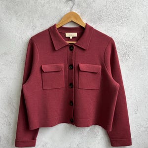 May include: A dark red button-down knit cardigan featuring a classic collar, long sleeves with cuffs, and two front chest pockets with flaps. The garment is secured with five dark brown buttons down the center and includes a small interior neck label.