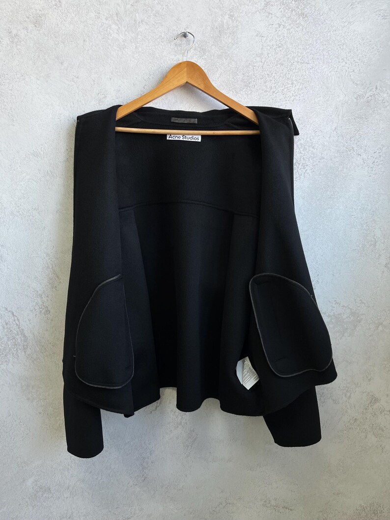 Pode incluir: Casaco preto pendurado num cabide de madeira. O casaco tem dois bolsos grandes e a etiqueta "Acne Studios" &eacute; vis&iacute;vel. O casaco &eacute; feito de um material espesso e tem um design simples e moderno.