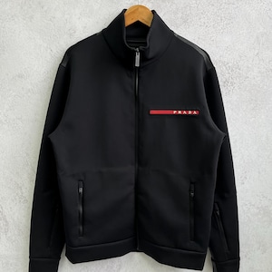 May include: Black zip-up jacket with a high collar and the brand name "PRADA" in red and white on the chest. The jacket has two side pockets and a full-length zip. The jacket is hanging on a wooden hanger.